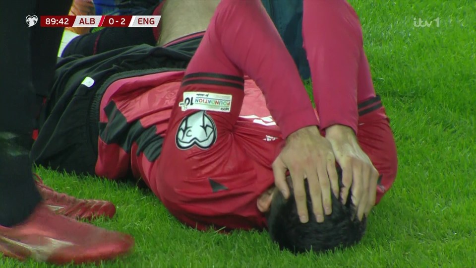 A soccer player in a red jersey lies on the grass with his hands on his head, indicating injury or distress.
